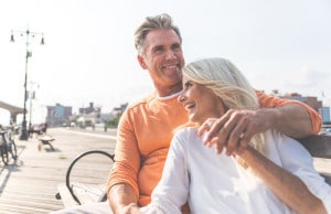 Man and woman sitting together on a beach boardwalk. Both are smiling. They are holding hands.