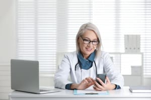 Mature female doctor with smartphone at table in office